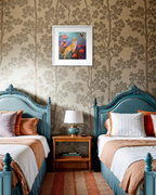 A styled bedroom interior with two teal beds, patterned wallpaper, a wooden nightstand with a lamp, and a framed colorful painting hanging on the wall.