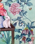 A decorative art print or wallpaper design featuring a white rabbit sitting on a wooden table next to pink peonies, with blue-green foliage, a butterfly, and additional rabbits on a light aqua background.