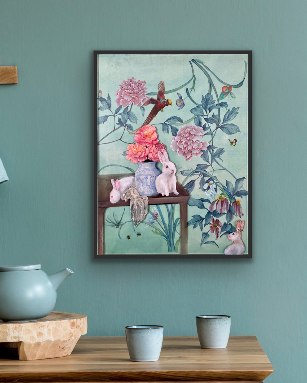 A framed art print featuring a whimsical floral still life with pink flowers, rabbits, and a bird, displayed on a teal wall above a wooden table with ceramic cups and a teapot.