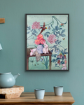 A framed art print featuring a whimsical floral still life with pink flowers, rabbits, and a bird, displayed on a teal wall above a wooden table with ceramic cups and a teapot.