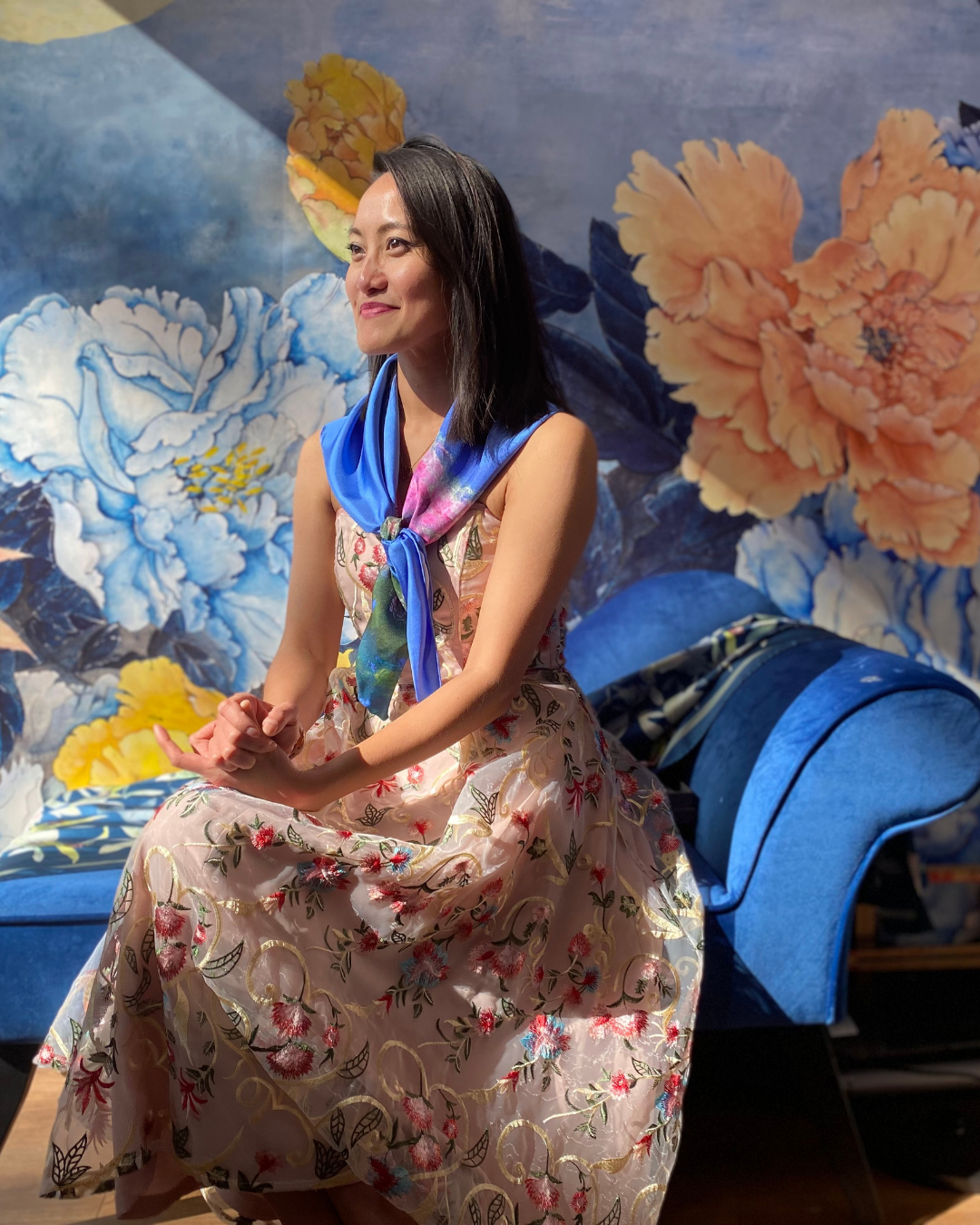 A woman sits on a blue velvet sofa in front of a large floral wall mural. She is wearing a sleeveless beige dress with a pink and green floral pattern and a bright blue scarf tied around her neck.
