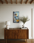 A vertical floral art print with blue, yellow, and white flowers hangs on a light beige wall above a mid-century modern wooden sideboard. The room is styled with a white table lamp on the left and a white ceramic vase filled with dried branches on the right.