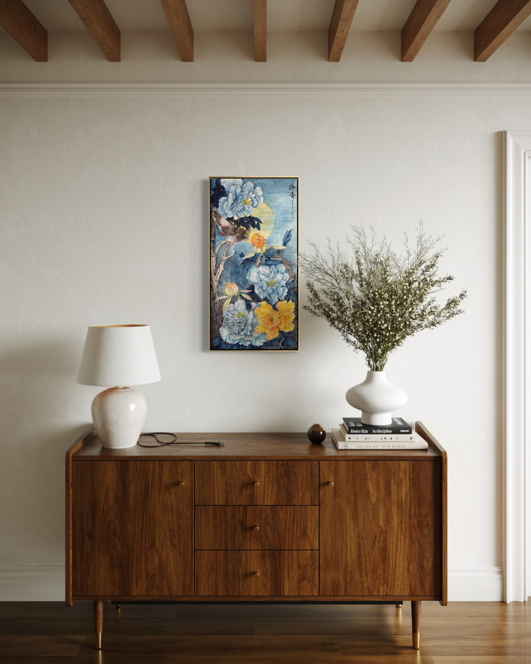 A vertical floral art print with blue, yellow, and white flowers hangs on a light beige wall above a mid-century modern wooden sideboard. The room is styled with a white table lamp on the left and a white ceramic vase filled with dried branches on the right.