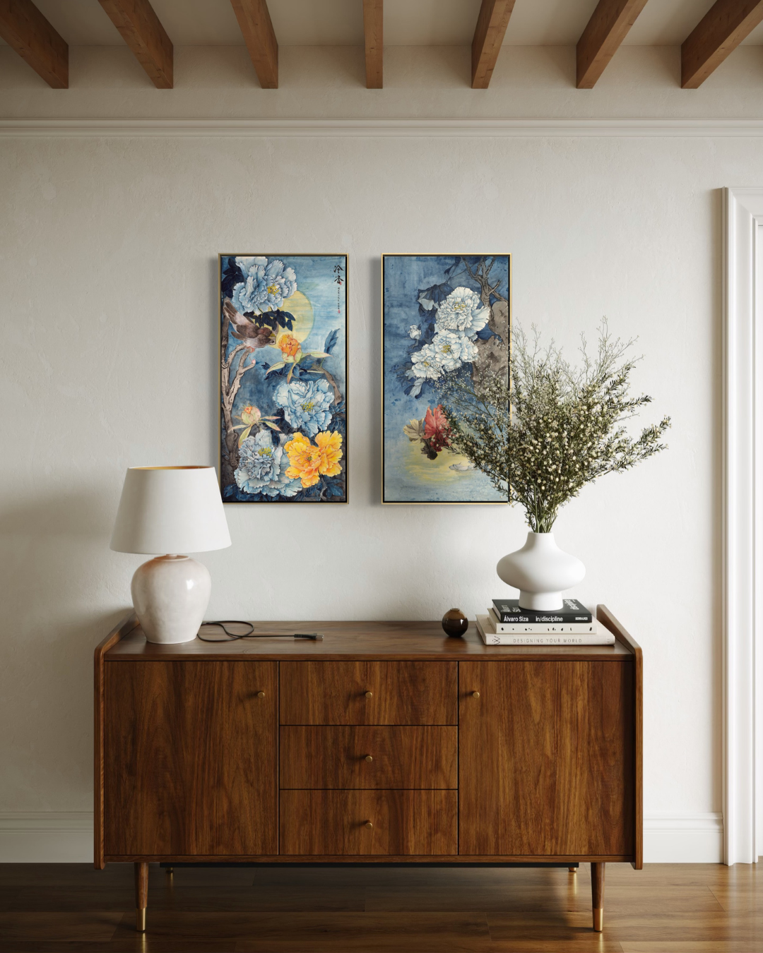 Two vertical framed floral art prints with blue, yellow, and white tones hang on a cream wall above a mid-century modern wooden sideboard. A white table lamp sits on the left, and a white vase with dried branches and books sits on the right.