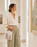 A woman in a cream blouse and sage green trousers carries a white embroidered handbag with a pearl strap in a bright art gallery.