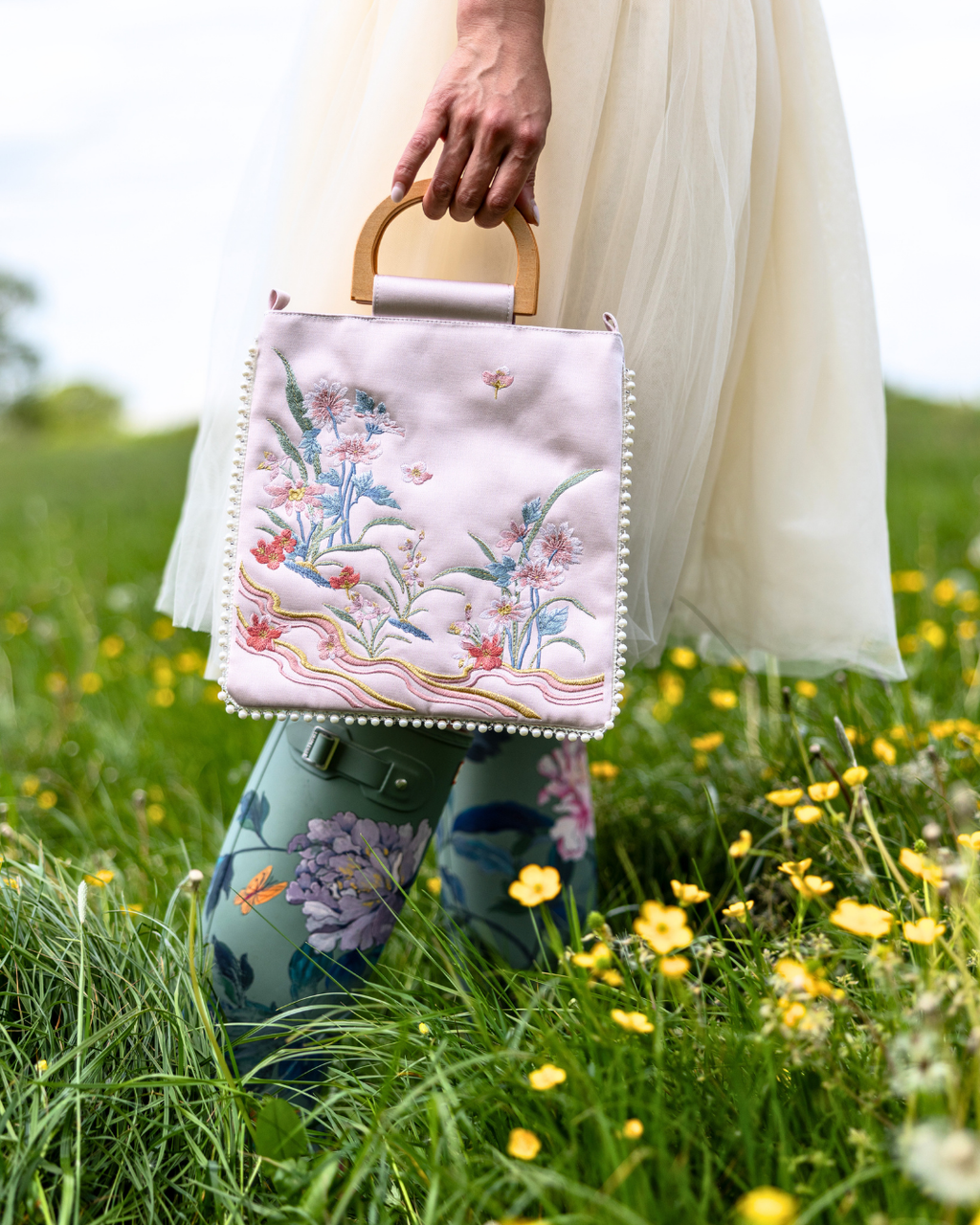A person in a white dress and floral rain boots holds a light pink embroidered handbag with a wooden top handle in a grassy field with yellow flowers.