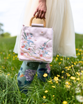 A person in a white dress and floral rain boots holds a light pink embroidered handbag with a wooden top handle in a grassy field with yellow flowers.