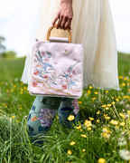 A person in a white dress and floral rain boots holds a light pink embroidered handbag with a wooden top handle in a grassy field with yellow flowers.