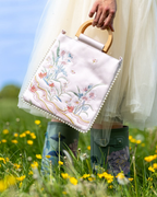 A person in a white tulle dress and green rain boots holds a square white embroidered handbag with a light wood top handle and pearl trim in a grassy field with yellow flowers.