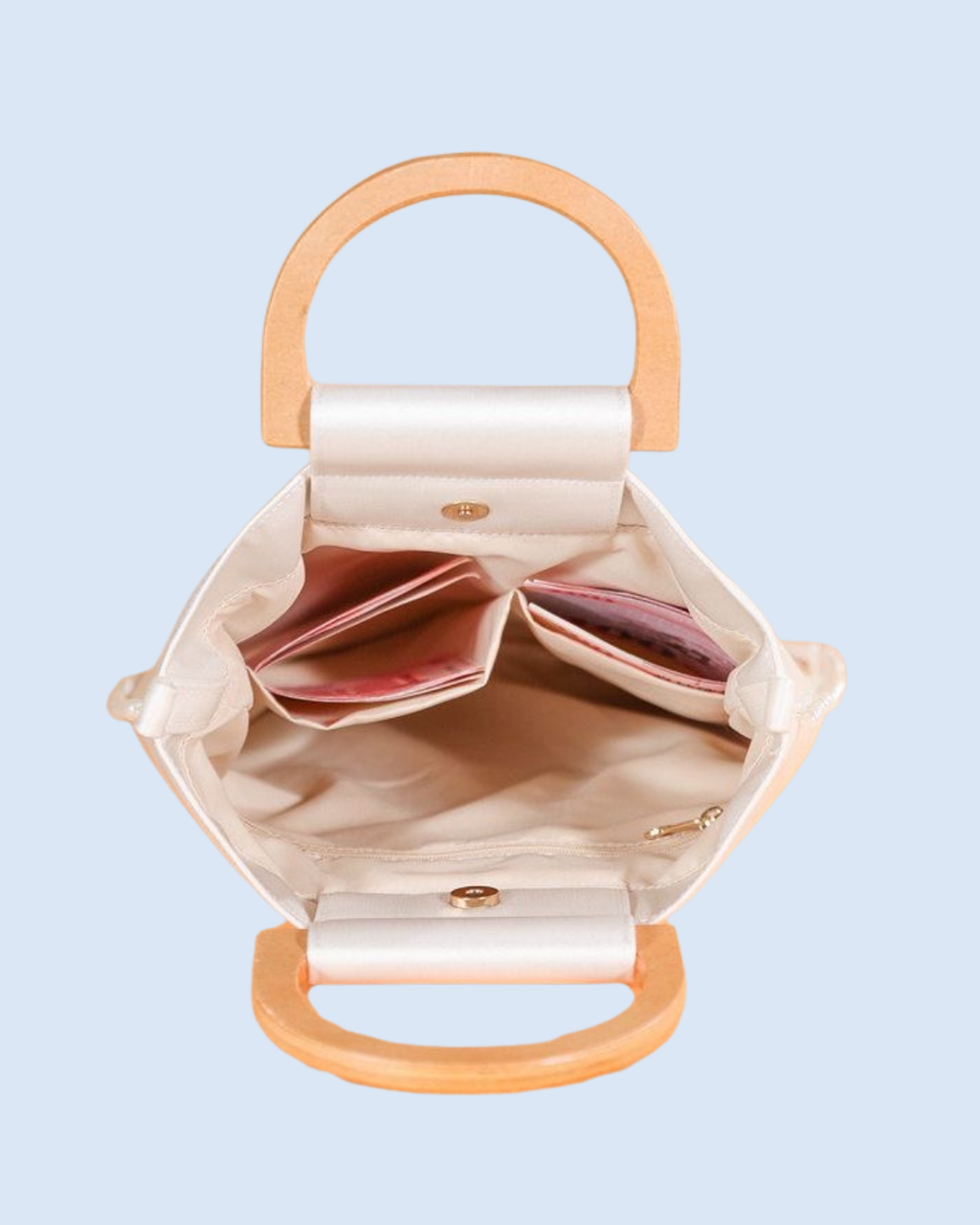 An open cream-colored fabric handbag with light wood handles, showing its interior compartments and pinkish-brown lining, set against a solid light blue background.