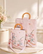 A light pink handbag set with floral embroidery, featuring a larger tote bag and a matching shoulder bag with light wood handles and pearl trim, displayed on a round table.