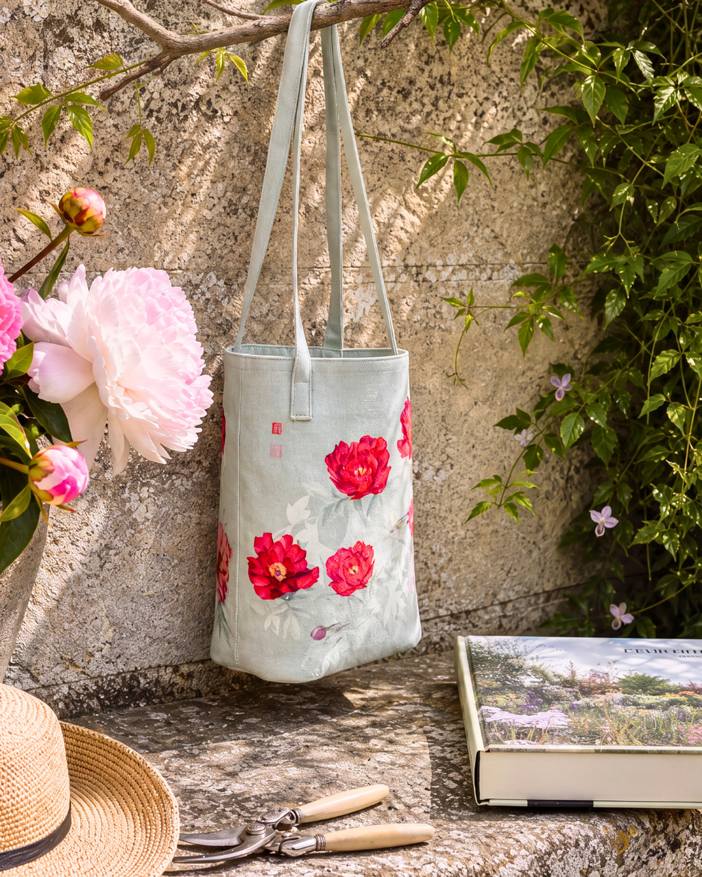 A light sage green fabric tote bag with red and pink floral prints hangs from a tree branch in a garden setting. The scene is styled with a straw hat, gardening shears, a book, and pink flowers against a textured stone wall.