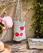A light sage green fabric tote bag with red and pink floral prints hangs from a tree branch in a garden setting. The scene is styled with a straw hat, gardening shears, a book, and pink flowers against a textured stone wall.
