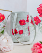 A light sage green fabric bucket bag with a floral print of red peonies and green leaves, featuring dual shoulder straps, styled with pink flowers in the foreground.