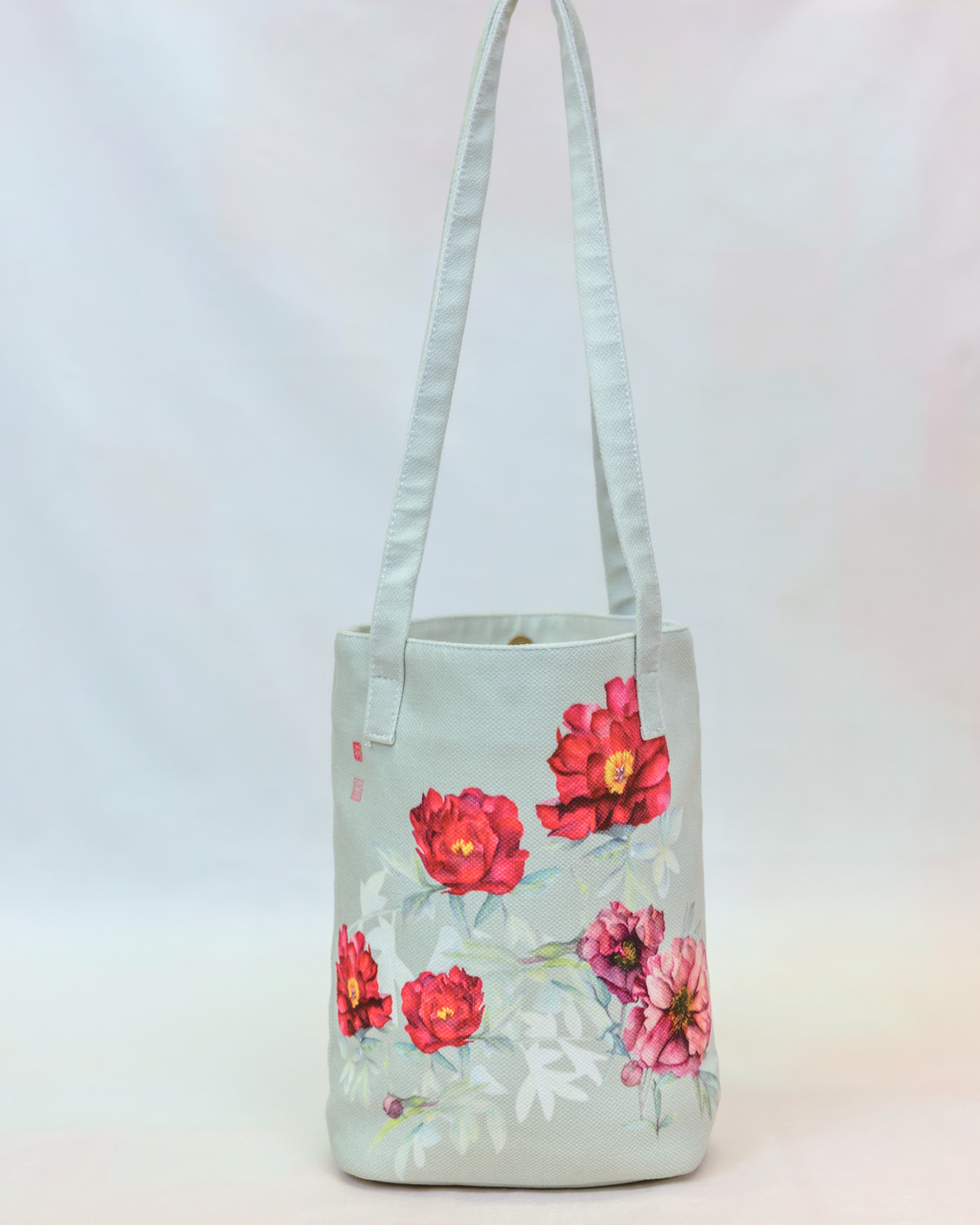 A light sage green canvas tote bag with long shoulder straps, decorated with a watercolor floral print of red and pink peonies with green leaves, set against a plain white background.