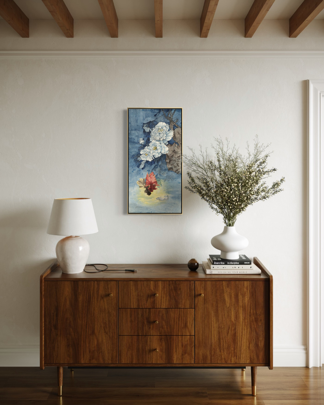 A framed vertical art print featuring a blue-toned floral painting with white blossoms and a small red accent, displayed on a cream wall above a mid-century modern wooden sideboard. The room includes a white table lamp, books, and a white vase with dried branches.