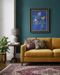 A framed floral art print hangs on a deep teal wall above a mustard yellow sofa with two patterned throw pillows. A wooden side table with a floral lamp and books sits to the left, next to a potted plant, and a patterned rug covers the floor.