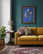 A framed floral art print hangs on a deep teal wall above a mustard yellow sofa with two patterned throw pillows. A wooden side table with a floral lamp and books sits to the left, next to a potted plant, and a patterned rug covers the floor.