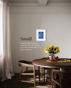 A styled interior scene showing a small framed art print on a beige wall above a round wooden dining table with chairs and a vase of yellow flowers. Text on the wall reads 'Small' with sizing/lifestyle image details.