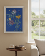 A framed botanical art print featuring blue, yellow, and purple flowers on a deep blue background, hanging on a beige wall next to a window with a garden view. A small wooden side table below holds a teacup and books, with part of a cream armchair visible.