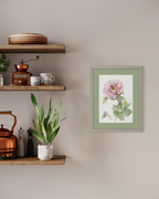 A framed botanical art print featuring a pink flower and green leaves, displayed on a beige wall next to wooden floating shelves with home decor.