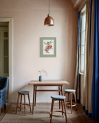 A styled interior scene with a small wooden table, three wooden stools with blue-gray cushions, a copper pendant light, beige walls, blue curtains, and a framed floral print on the wall.
