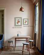 A styled interior room showing two framed botanical art prints on a beige wall above a small wooden table and stools. The prints are labeled as large 29.