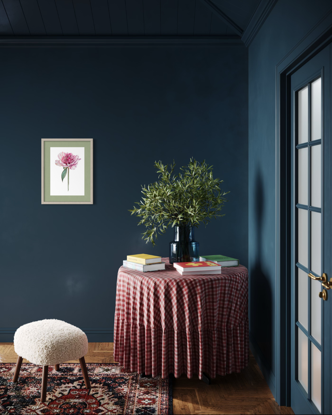 A styled interior corner with dark blue walls, a small round table covered in a red-and-white gingham tablecloth, a black vase with green foliage, books, and a framed floral print on the wall. A white boucle stool and patterned rug are also visible.