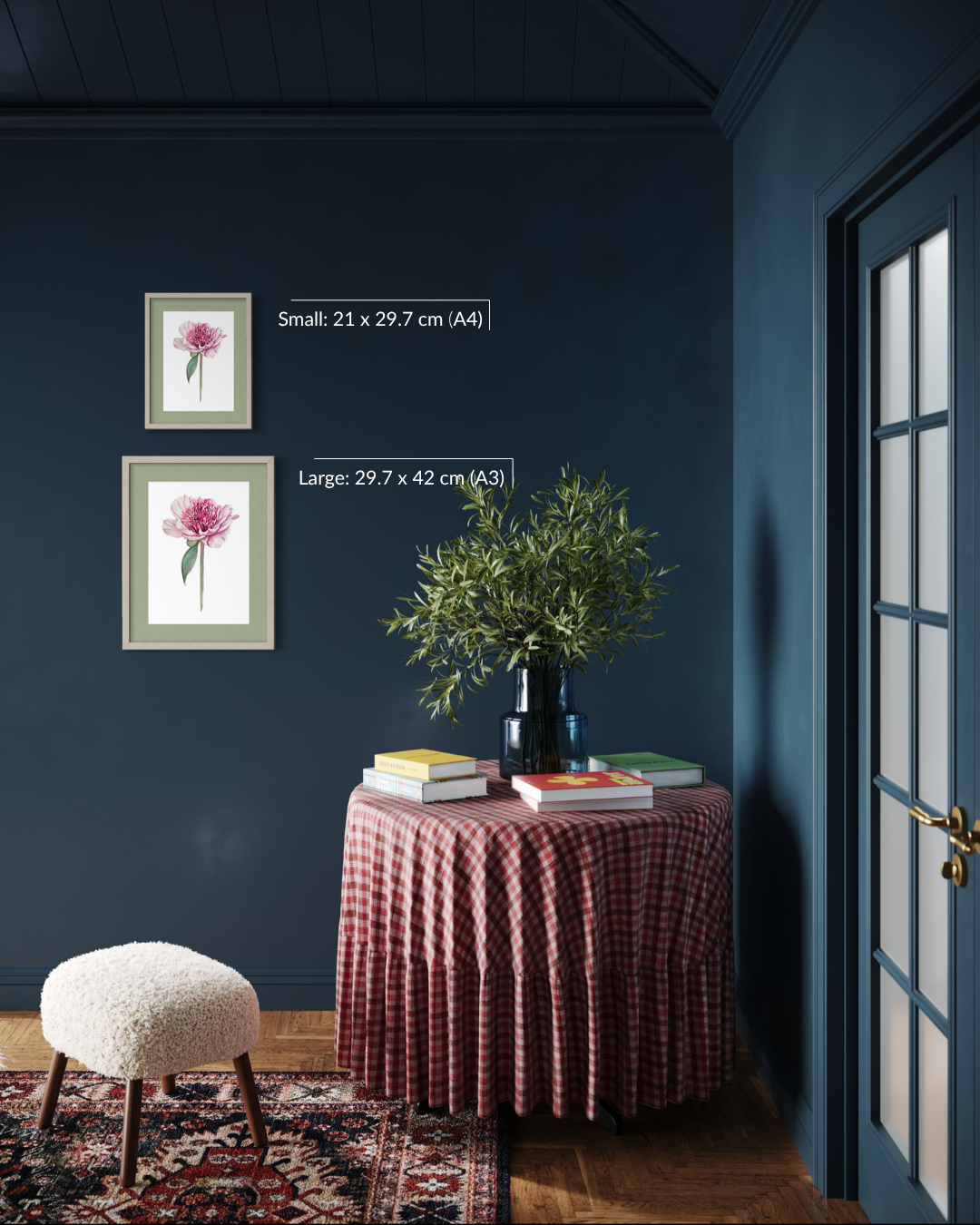 A styled interior scene showing two framed art prints on a dark blue wall above a small table with books and a plant. White text labels indicate sizes: Small 21 x 29.