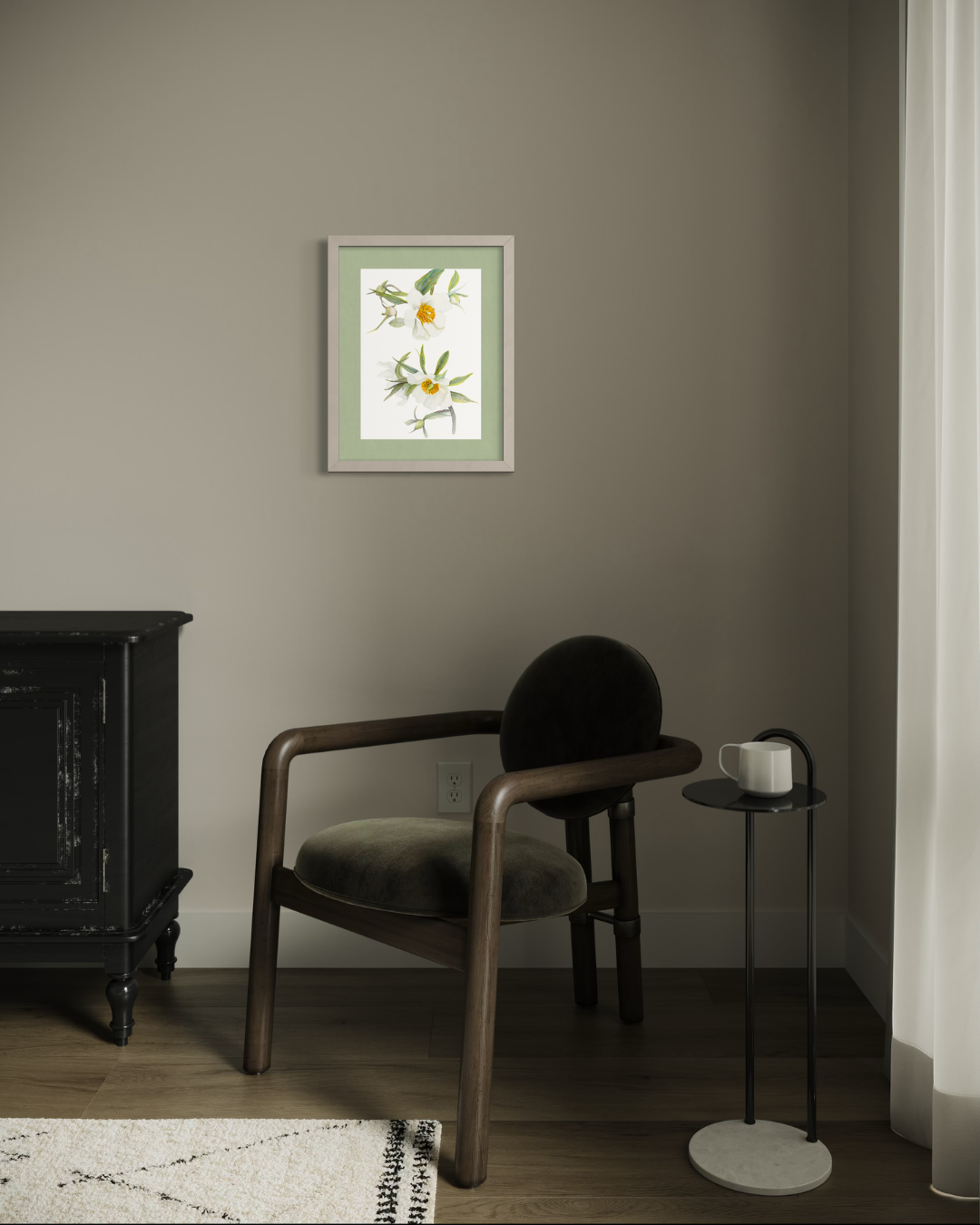 A framed botanical art print hangs on a taupe-gray wall in a styled room setting with a dark wood armchair, a black side table with a white mug, and a dark cabinet.