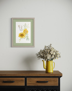 Framed botanical wall art featuring yellow flowers and green leaves, displayed on a light gray wall above a wooden console table with a yellow pitcher of dried flowers.
