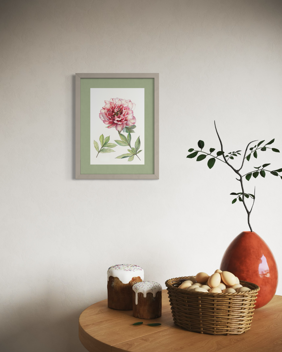 A framed botanical art print of a pink peony with green leaves, displayed on a neutral wall above a wooden table styled with a wicker basket, small jars, and a terracotta vase.