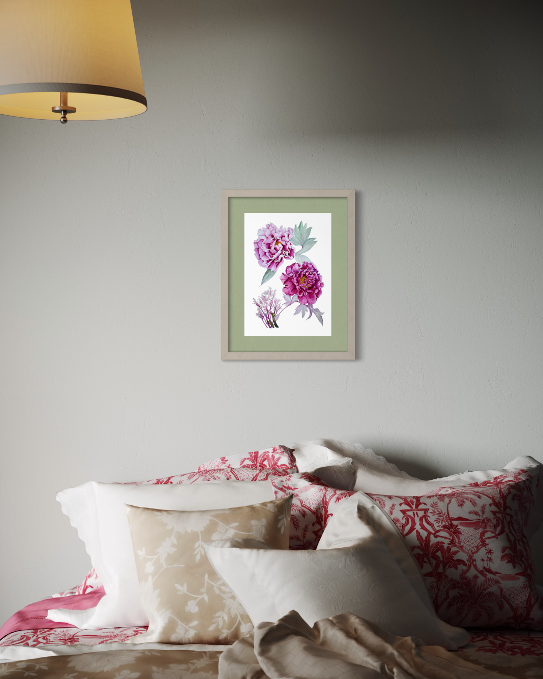 A framed botanical art print featuring pink peonies with green leaves, displayed on a light gray wall above a bed with patterned pink and beige pillows and a lit lamp in the corner.