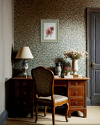 A styled interior corner featuring a dark wood writing desk with a matching upholstered chair, a table lamp, ceramic vessels, and a framed floral print on a patterned wallpapered wall.
