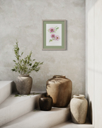 A framed botanical art print featuring pink flowers and green leaves, styled on a textured light gray wall above rustic ceramic vases on a stone staircase.