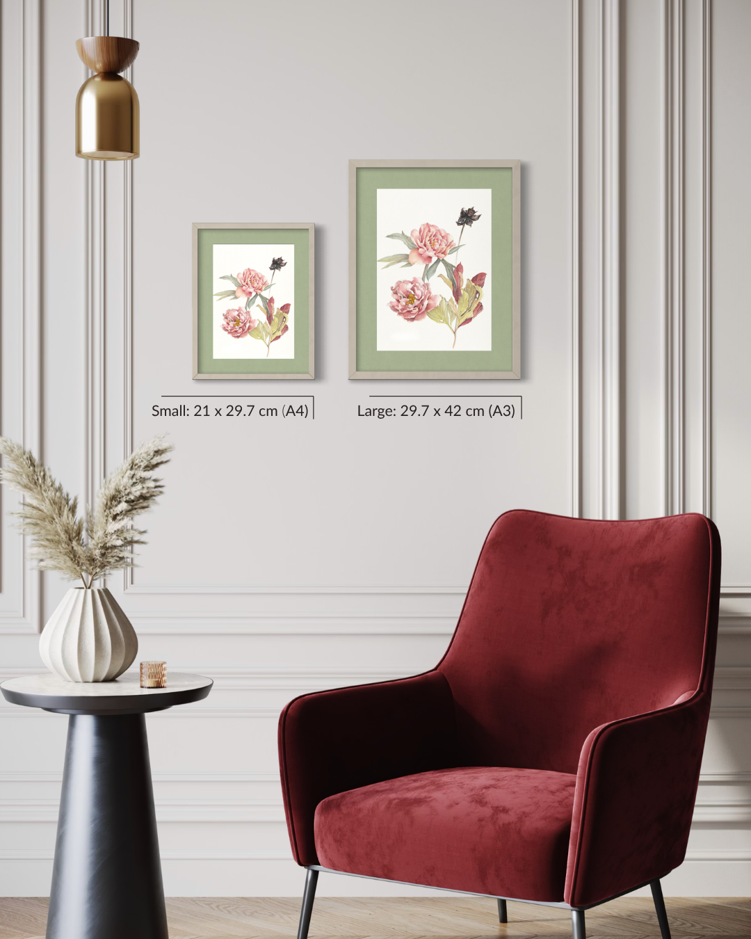 Two framed botanical art prints with pink flowers are displayed on a light gray paneled wall above a deep red velvet armchair and a small side table with a vase of dried grass.