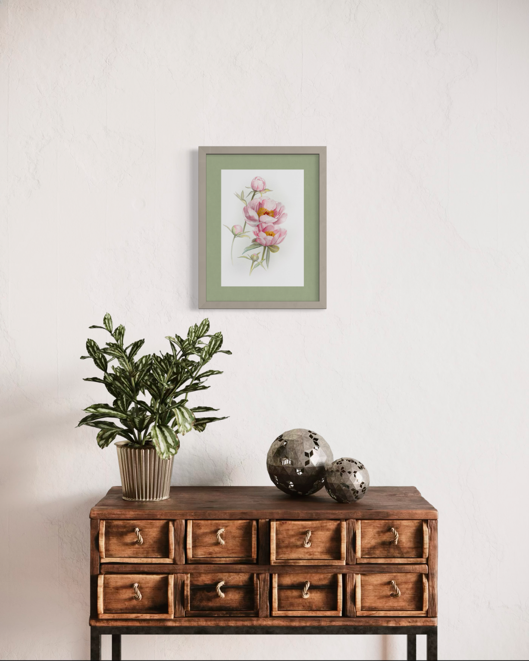 A framed botanical art print of pink flowers with a sage green border and gray frame, displayed on a textured white wall above a rustic wooden console table with a potted plant and decorative spheres.