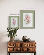 Two framed botanical art prints of pink flowers are displayed on a white wall above a wooden cabinet, with size labels 'Small: 21 x 29.7 cm (A4)' and 'Large: 29.