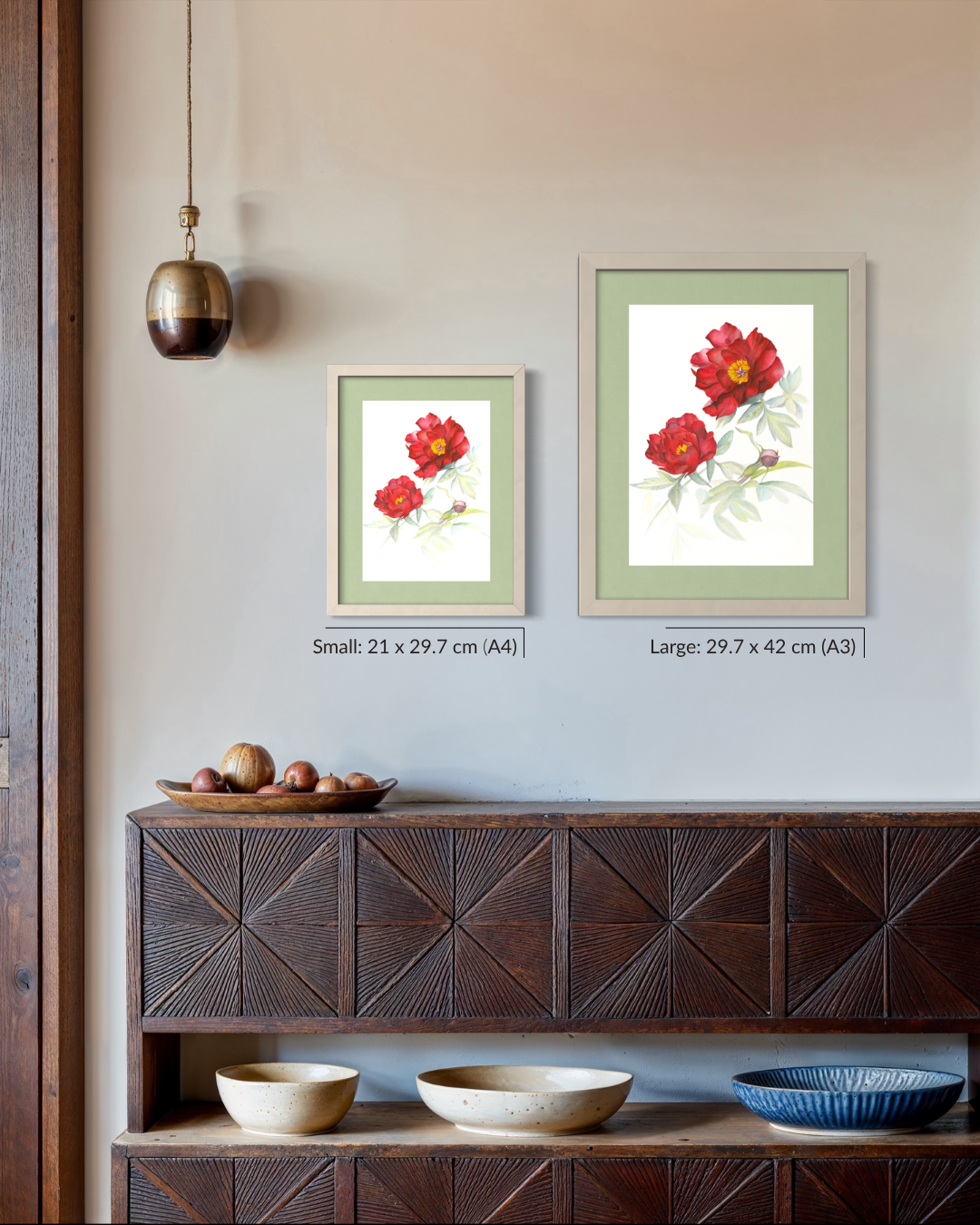 Two framed floral art prints hang on a beige wall above a dark wood console table. The prints show red flowers with green leaves on white backgrounds, one small and one large, with size labels beneath them.