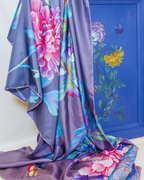 A purple silk scarf with a vibrant floral print featuring pink, blue, and yellow flowers, draped elegantly in front of a blue wooden panel decorated with painted flowers.