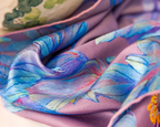 A close-up of a draped silk scarf featuring a watercolor-style floral pattern in shades of blue, purple, and pink over a dusty lavender base, with a glimpse of another patterned textile in the background.
