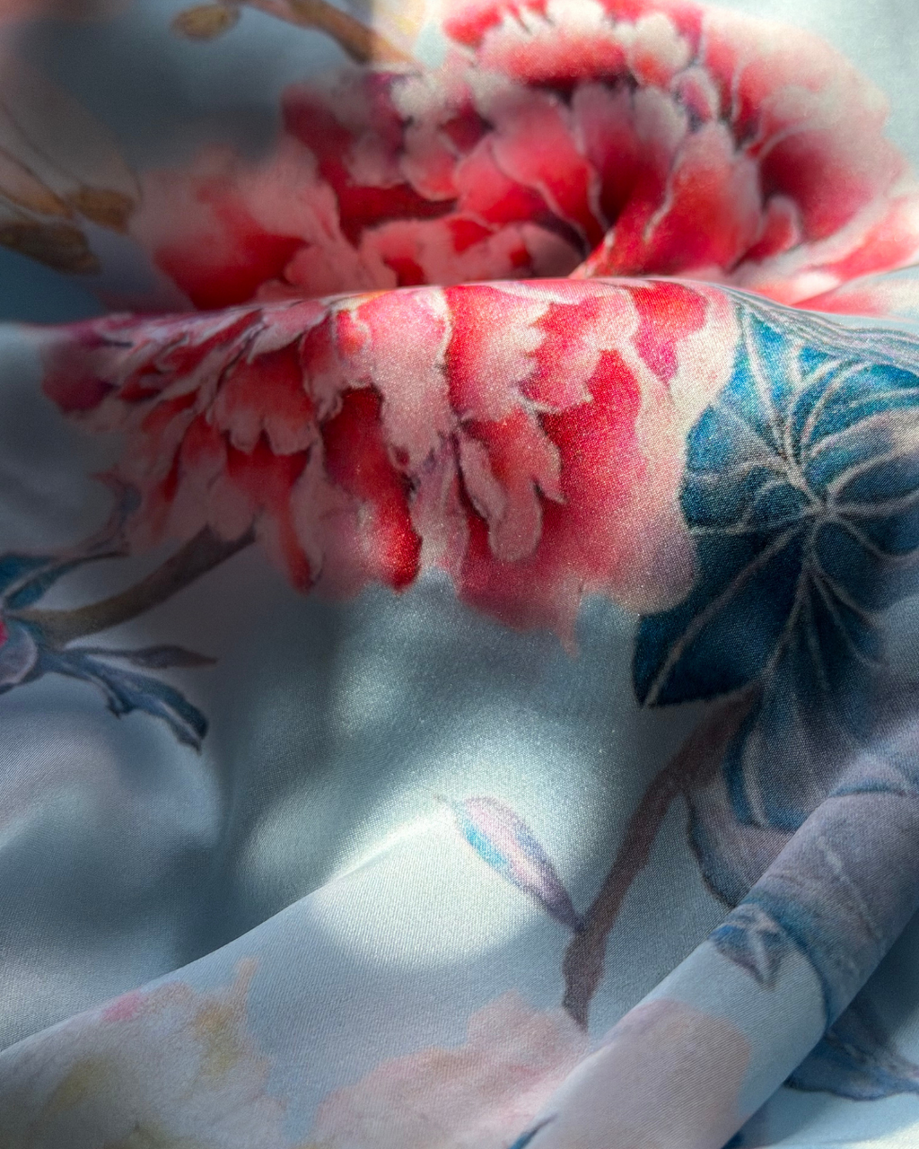 A close-up of a lightweight fabric with a watercolor-style floral print, featuring large pink and red blossoms and blue leaves on a pale blue background.