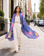 A woman walking on a city sidewalk wearing a flowing purple and blue floral scarf draped over her shoulders, paired with cream trousers and heels.