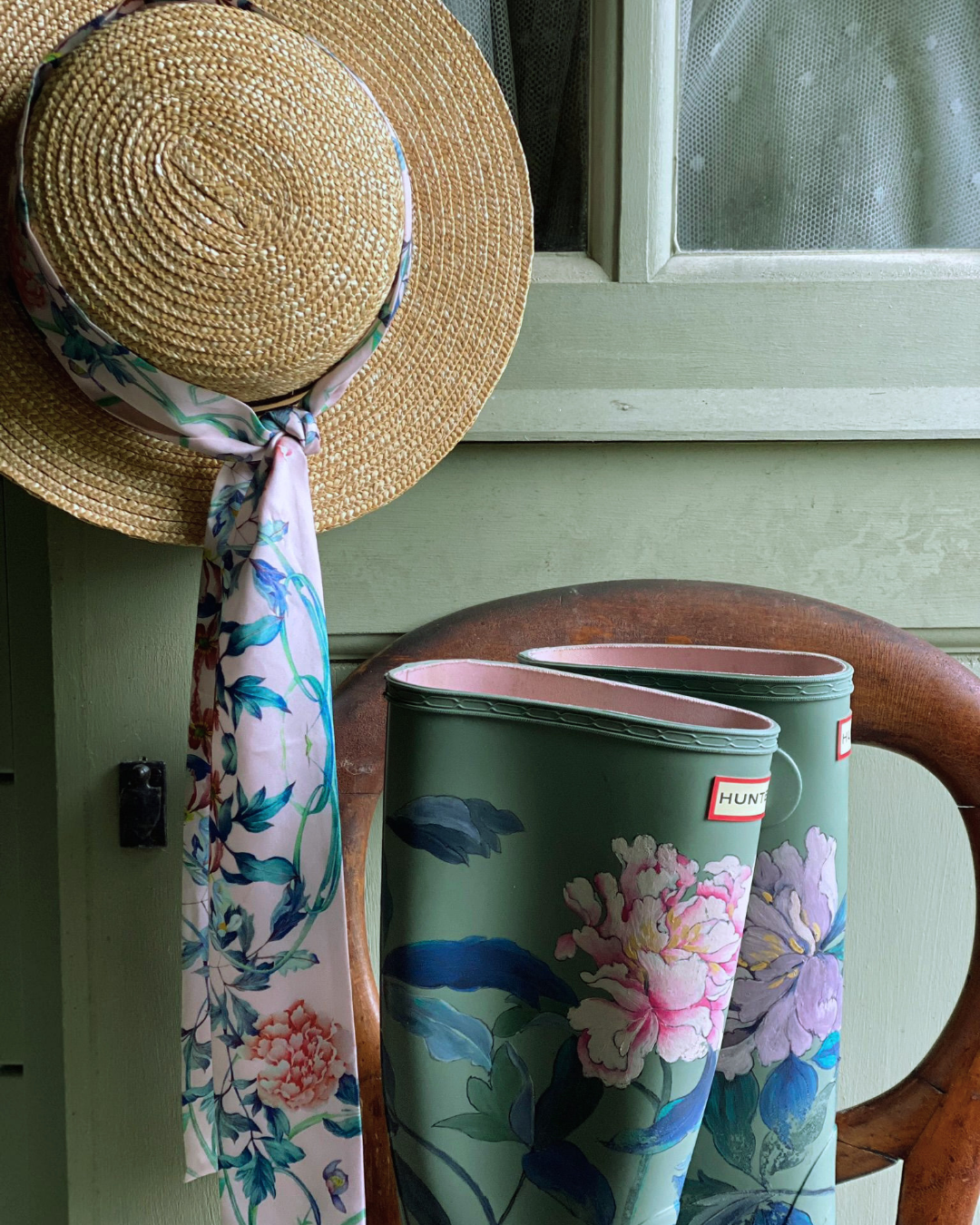 A pair of green Hunter rain boots with a pink and blue floral print, styled on a wooden chair next to a straw hat with a matching floral scarf.