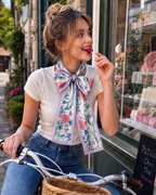 A woman wearing a white t-shirt and blue jeans sits on a wicker basket bicycle in front of a shop window, wearing a large floral bow scarf with pink, blue, and green patterns and lavender borders.