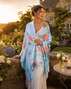 A woman stands in a sunlit garden wearing a light blue wrap or shawl with large pink and teal floral prints over a cream dress. She holds a glass of white wine, with flowers, a stone wall, and a cottage in the background.