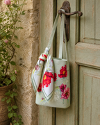 A fabric tote bag with a red and pink floral print hangs from a rustic metal door hook on a weathered green door, next to a potted plant.