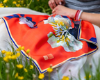 A close-up of a bright orange silk scarf with a large white and yellow floral print, blue border trim, and a small red seal, draped over a person's shoulder in an outdoor setting with yellow flowers.
