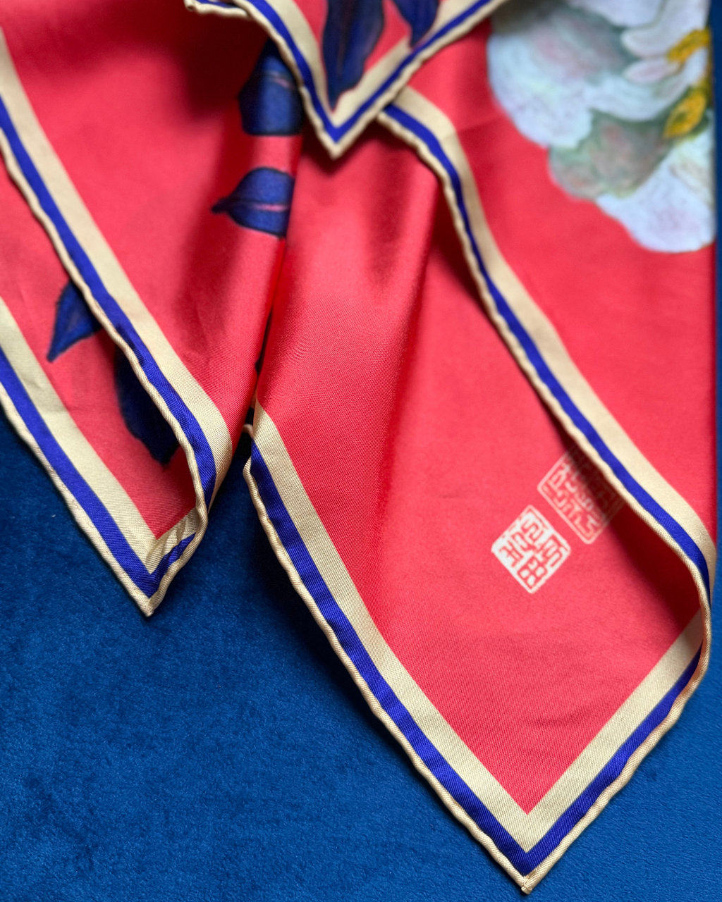 A close-up of a folded red silk scarf with blue and gold borders, featuring a white and yellow floral print in the top right corner and two red Chinese-style seals.