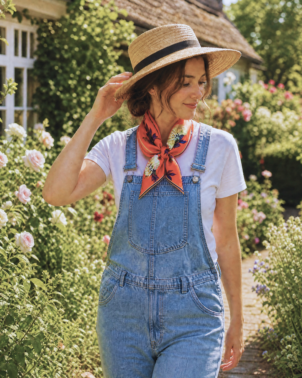A woman standing in a sunlit garden wearing light blue denim overalls over a white T-shirt, with a bright orange floral neck scarf and a straw hat with a black band.